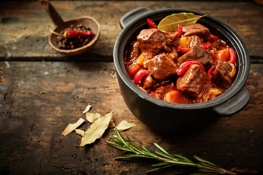 Pentola di gulash ungherese con peperoni rossi e foglie d’alloro su tavolo rustico.