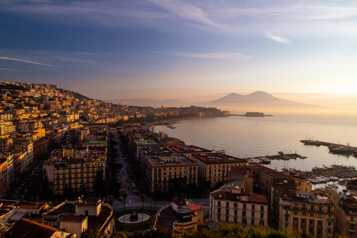 Vista panoramica del Golfo di Napoli al tramonto con il Vesuvio sullo sfondo, il Lungomare illuminato e il porto osservati dall’alto.
