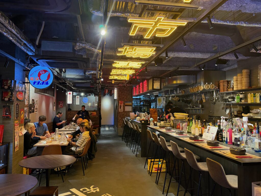 Interno del ristorante Moon & Back a Kyoto, con bancone, cucina a vista, tavoli e insegne luminose giapponesi.