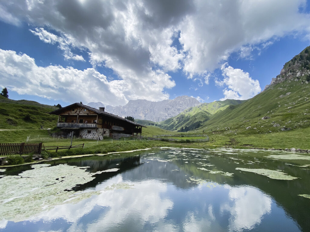 Baita di montagna accanto a un laghetto alpino circondato da prati e montagne