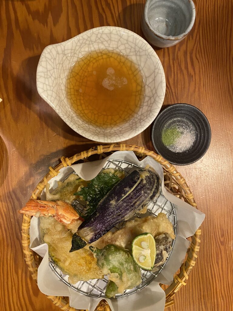 Tempura di gamberi e verdure, tra cui melanzana, servita in un cestino di bambù con salsa e condimenti.