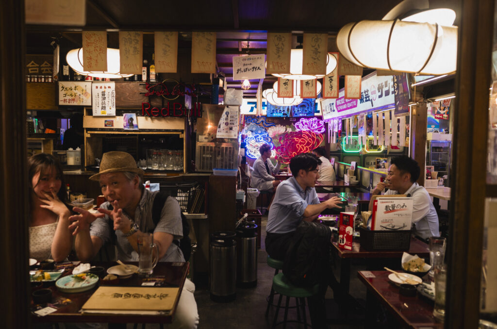 Interno di un izakaya giapponese con clienti seduti ai tavoli, lanterne illuminate e insegne al neon.